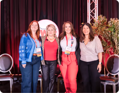 Four women standing side by side on stage with a red and black backdrop and empty chairs around them.