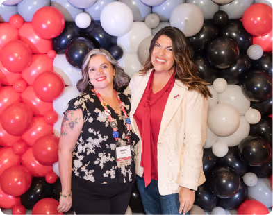 Two smiling women standing in front of a balloon wall with red, black, white, and lavender balloons.