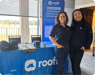Two women smiling standing by a Roofr branded table with hats and a tech display in a sunlit room.