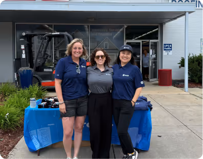 Three women smiling in front of a forklift and table with promotional items outside a building.
