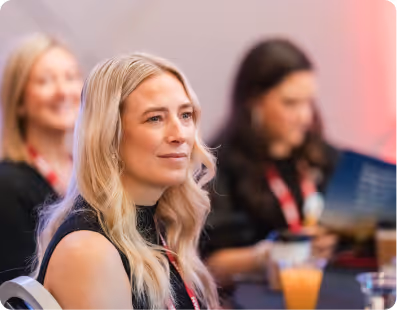 Blonde woman attentively listening at an event with two blurred women in the background.