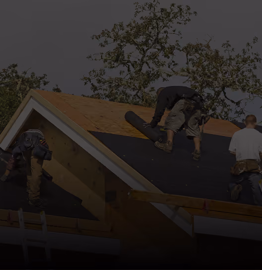 Construction worker wearing a safety harness carrying a plank on a rooftop with blue sky and clouds in the background.