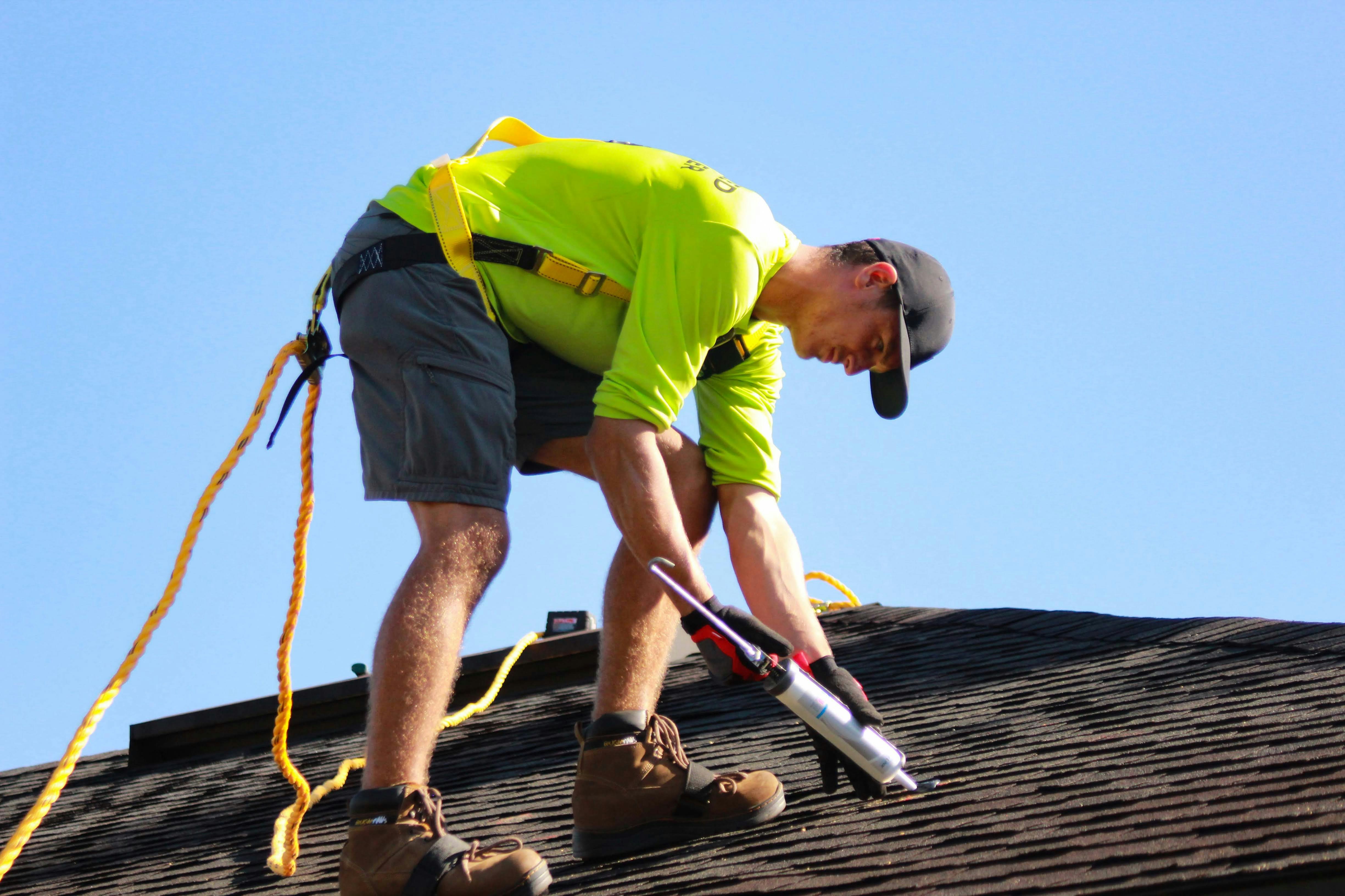 A roofer working hard on a roof, caulking asphalt tiles, in bright yellow shirt and attached with a safety harness.