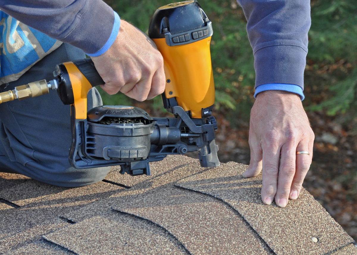 A close-up photo of a roofer using a nail gun to attach asphalt shingles to a roof ridge.