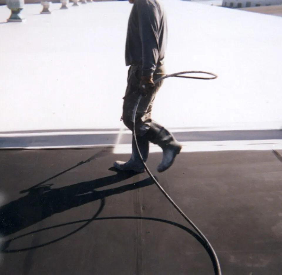 A roofing contractor on the roof wearing boots with a spray hose applying roof coating to a commercial flat roof.
