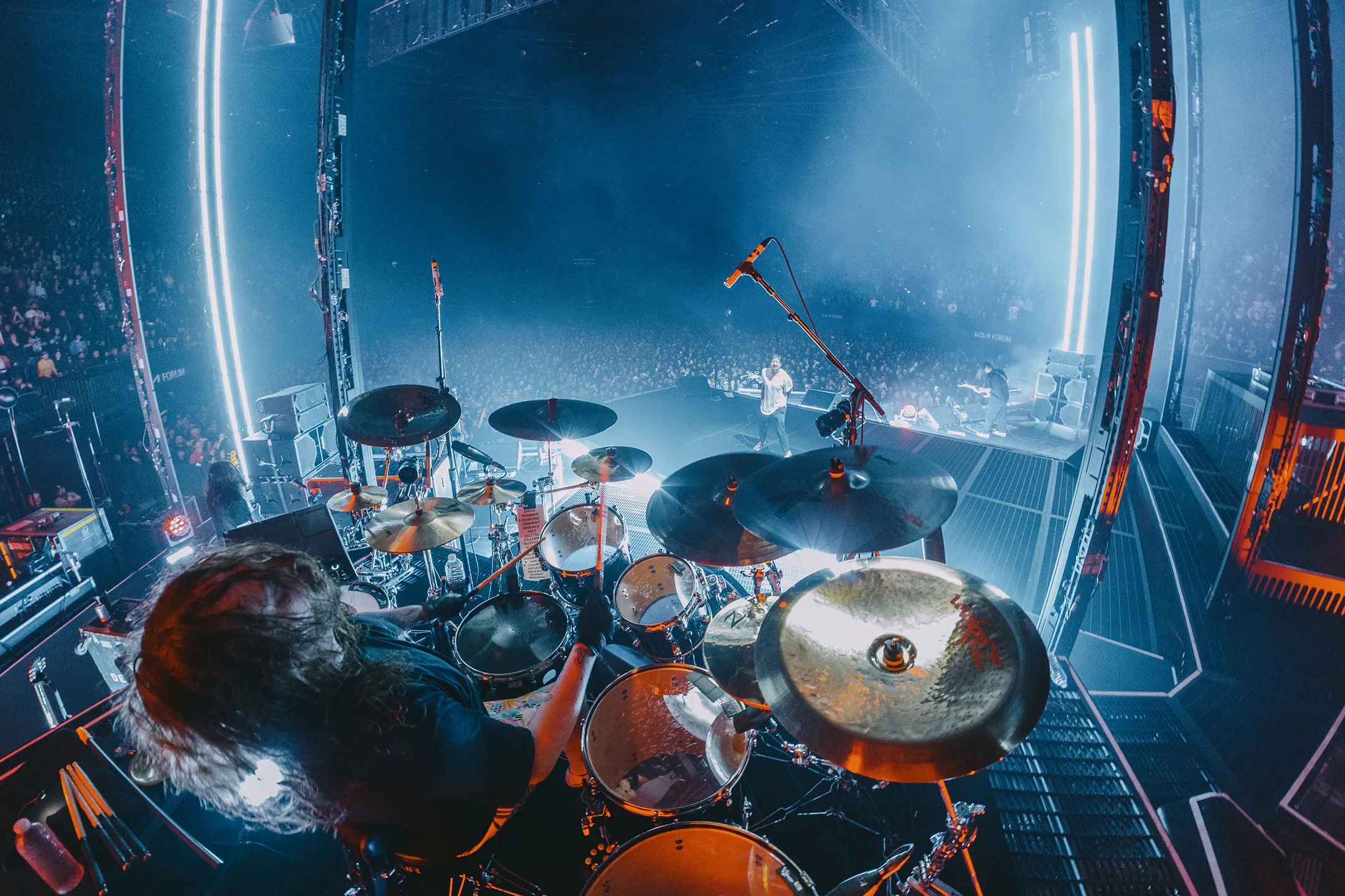 Deftones drummer Abe Cunningham playing his drum kit live on an elevated stage overlooking a packed arena.