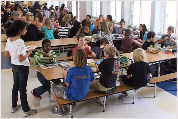 Crowded school cafeteria with children eating lunch at long tables.