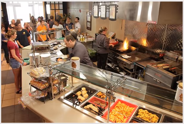 College dining hall with chefs cooking and students waiting in line for food.