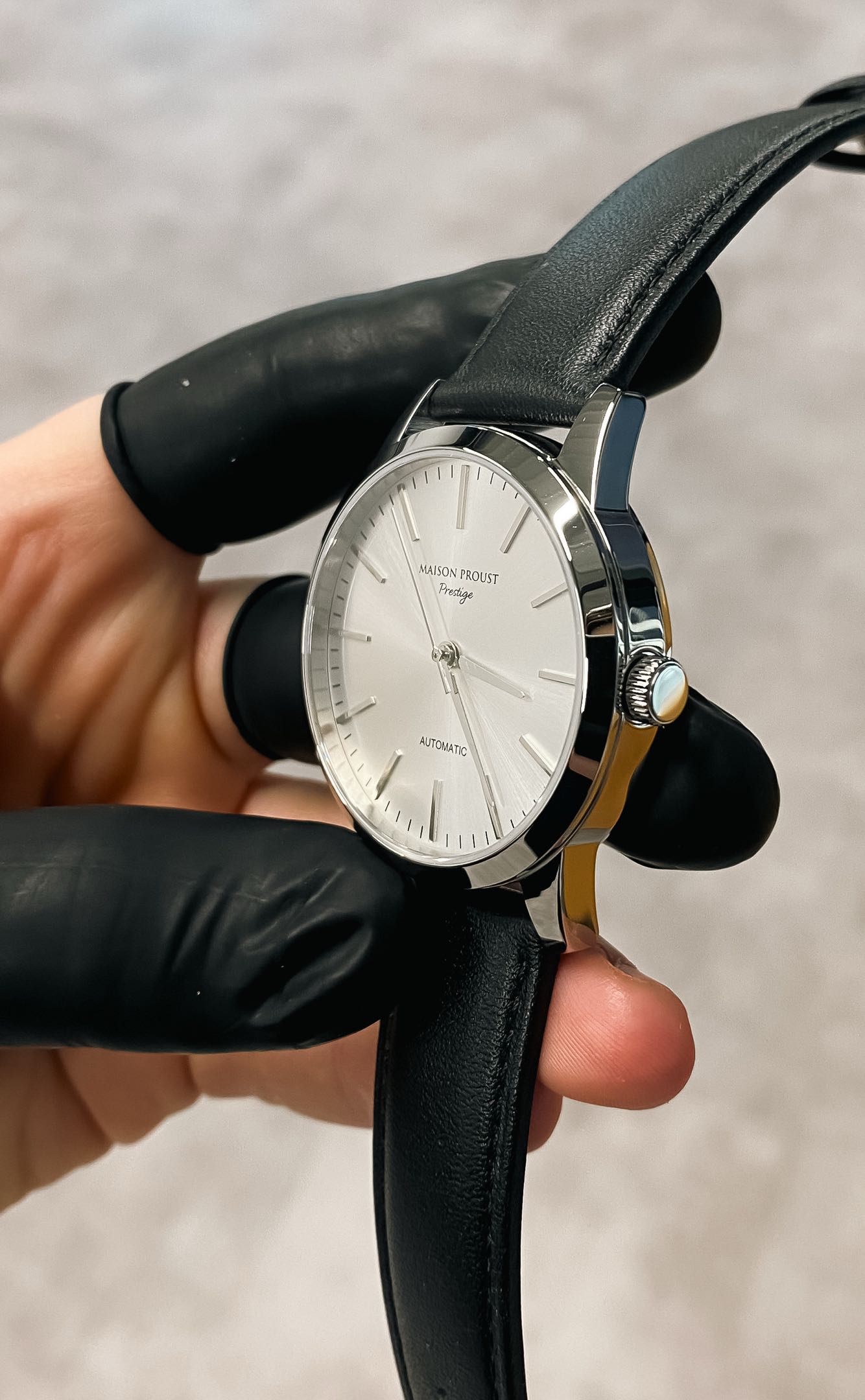 Close-up of a Maison Proust Prestige automatic wristwatch with a silver dial and black leather strap against a dark background.