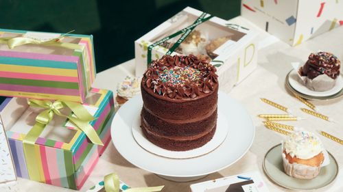 Pastel de chocolate de dos pisos con chispas de colores rodeado de cajas de regalo y cupcakes.
