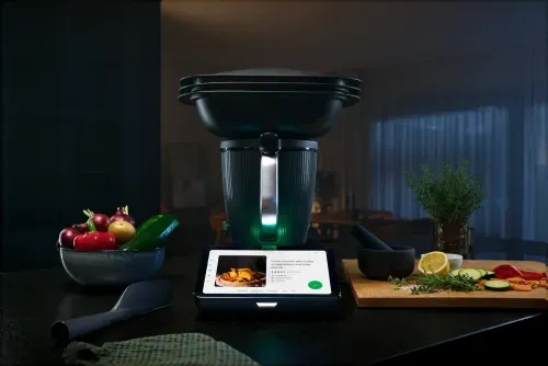 Robot de cocina con pantalla digital mostrando receta de pollo rostizado junto a verduras frescas y hierbas.
