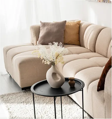 A curved, light-colored modular sofa with a black side table holding a modern white vase.