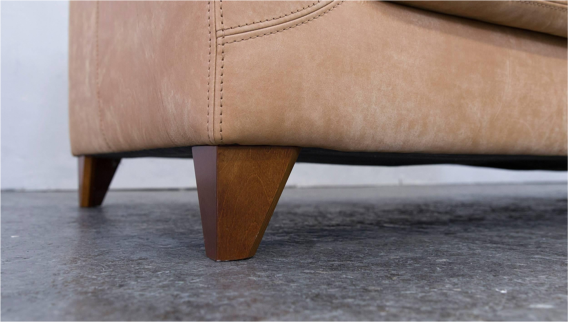 A close-up of a tan leather sofa's corner showing the stitching detail and a tapered wooden leg on a concrete floor.