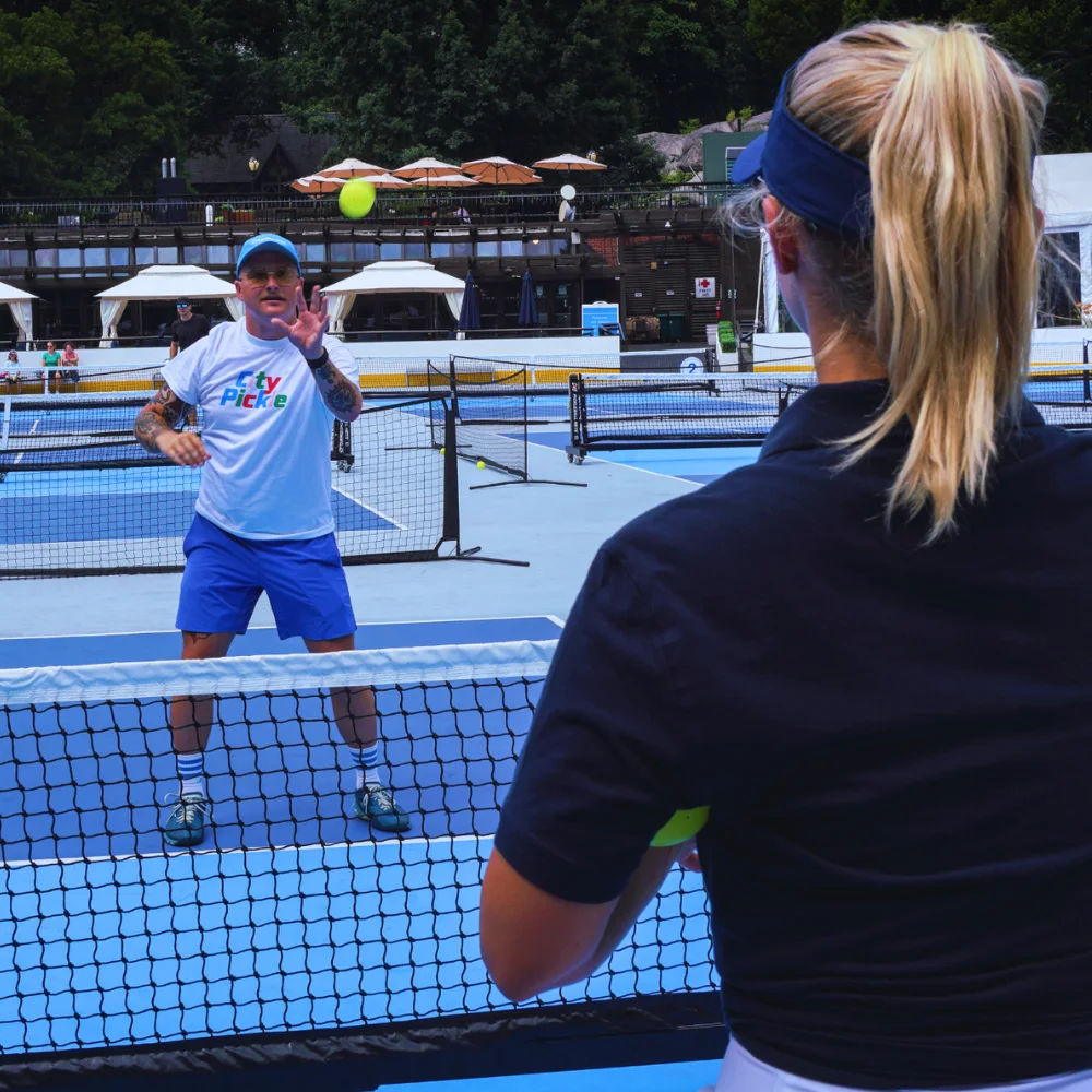 A coach feeding balls across the net to a player during a lesson on an outdoor court