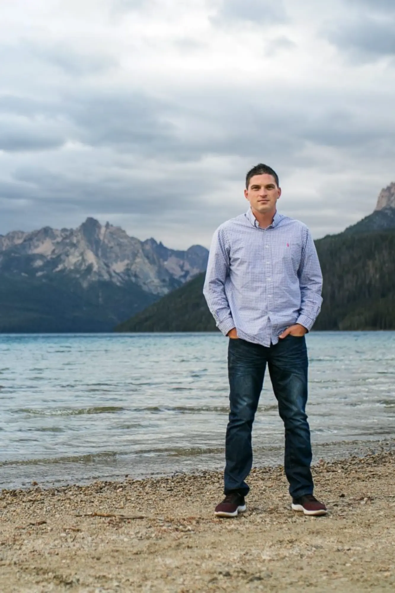 A man standing in front of a lake with mountains in the background.