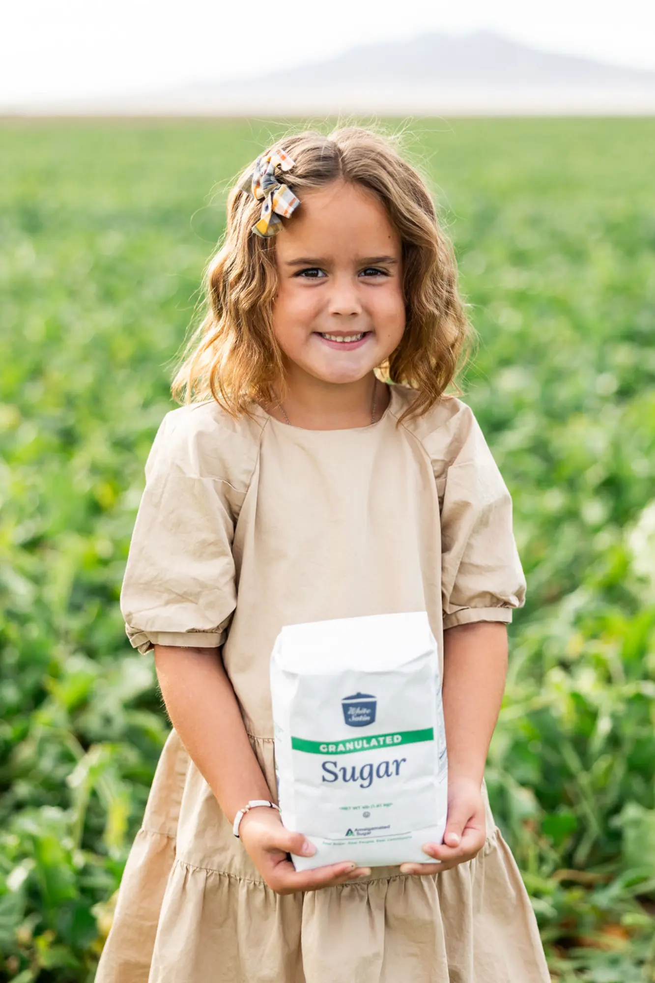 Smiling girl in a beige dress holding a bag of granulated sugar in a green field.
