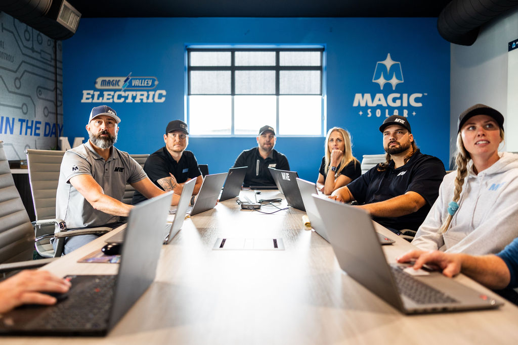 Group of six people in casual work attire sitting around a conference table with laptops in a modern office featuring blue walls with Magic Valley Electric and Magic Solar logos.