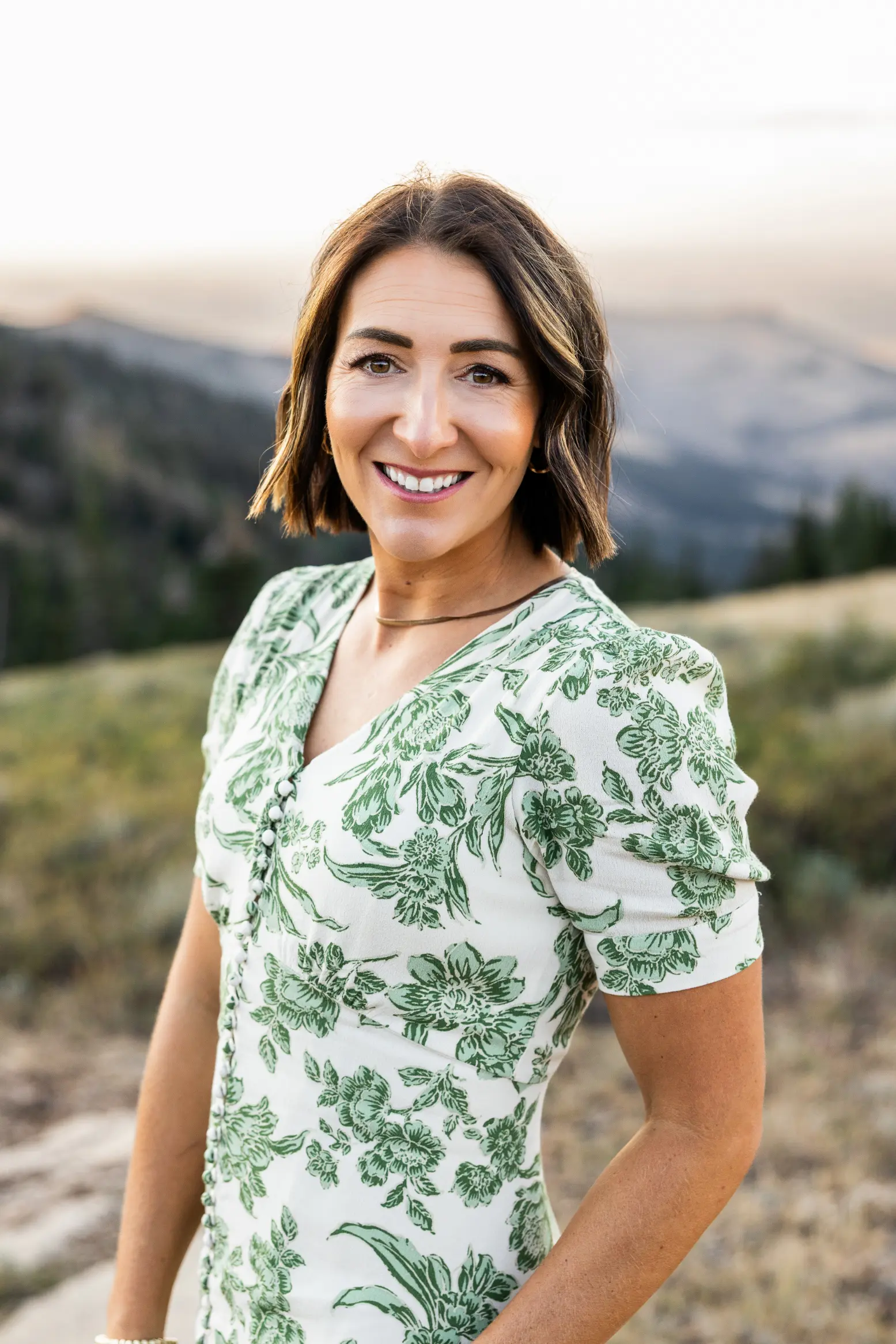 Smiling woman with short brown hair wearing a white dress with green floral patterns outdoors.