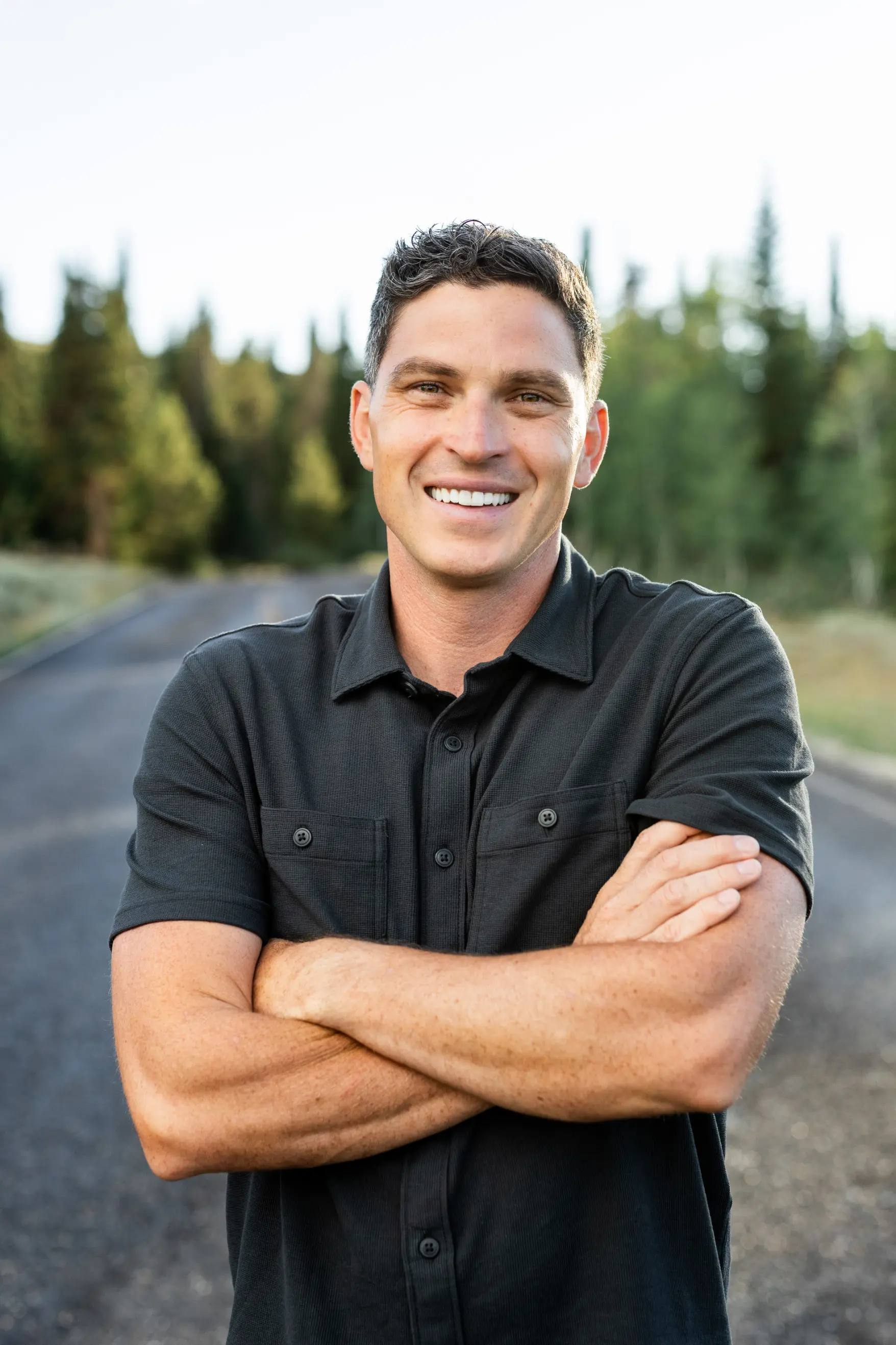 Smiling man in dark short-sleeve shirt with arms crossed standing on a road with trees behind him.