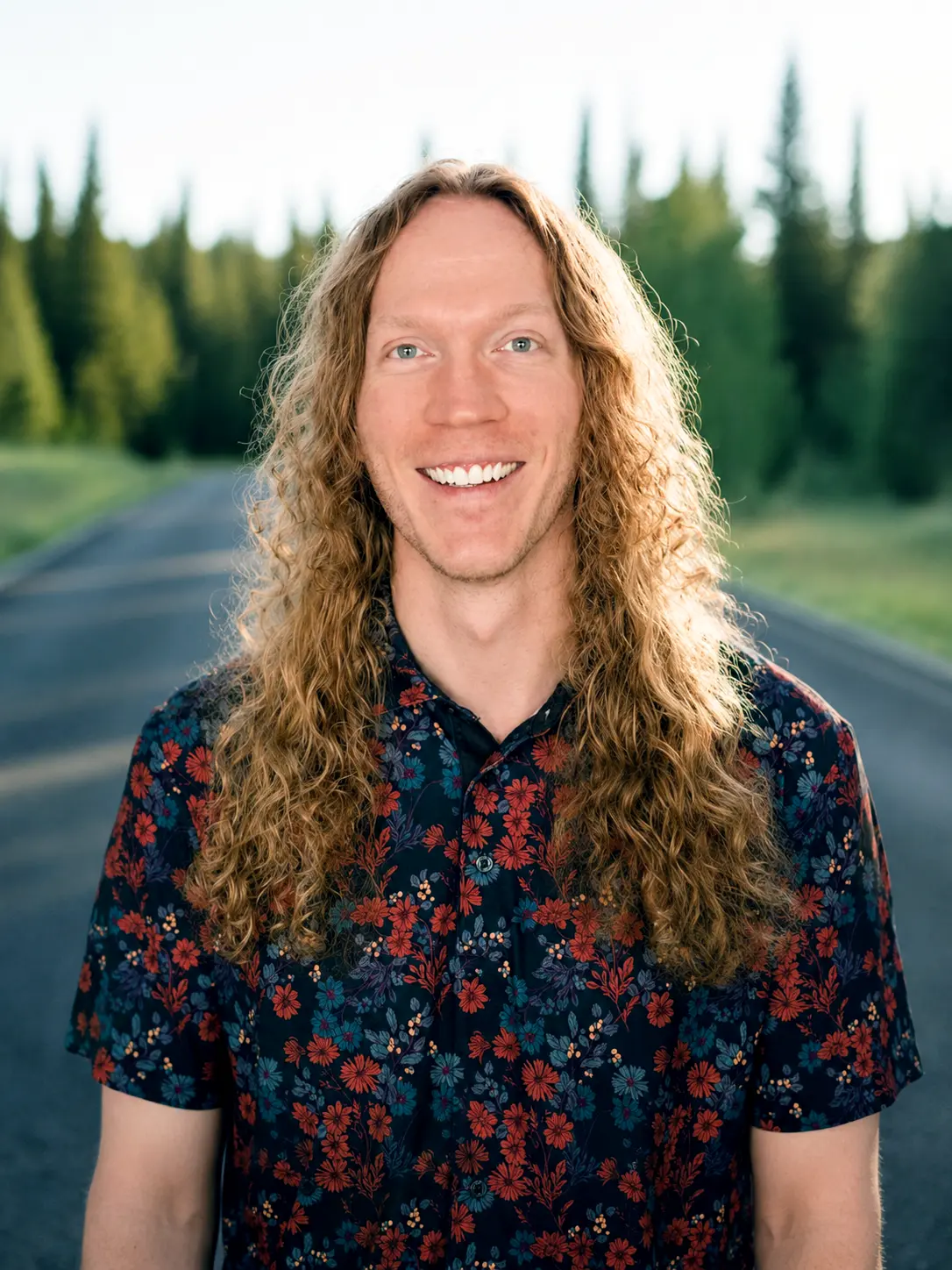 Smiling man with long curly hair wearing a floral shirt standing on a road with trees in the background.