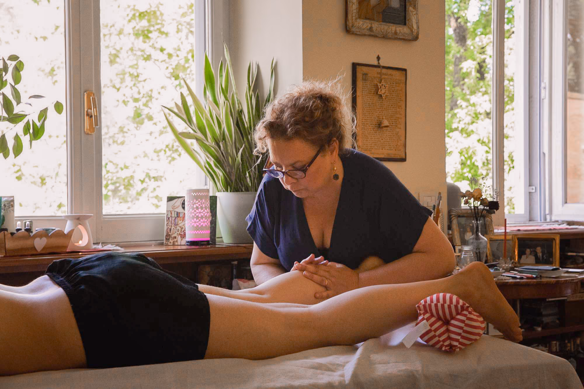 A woman in glasses gives a leg massage to a person lying face down on a massage table in a cozy room with plants and windows.