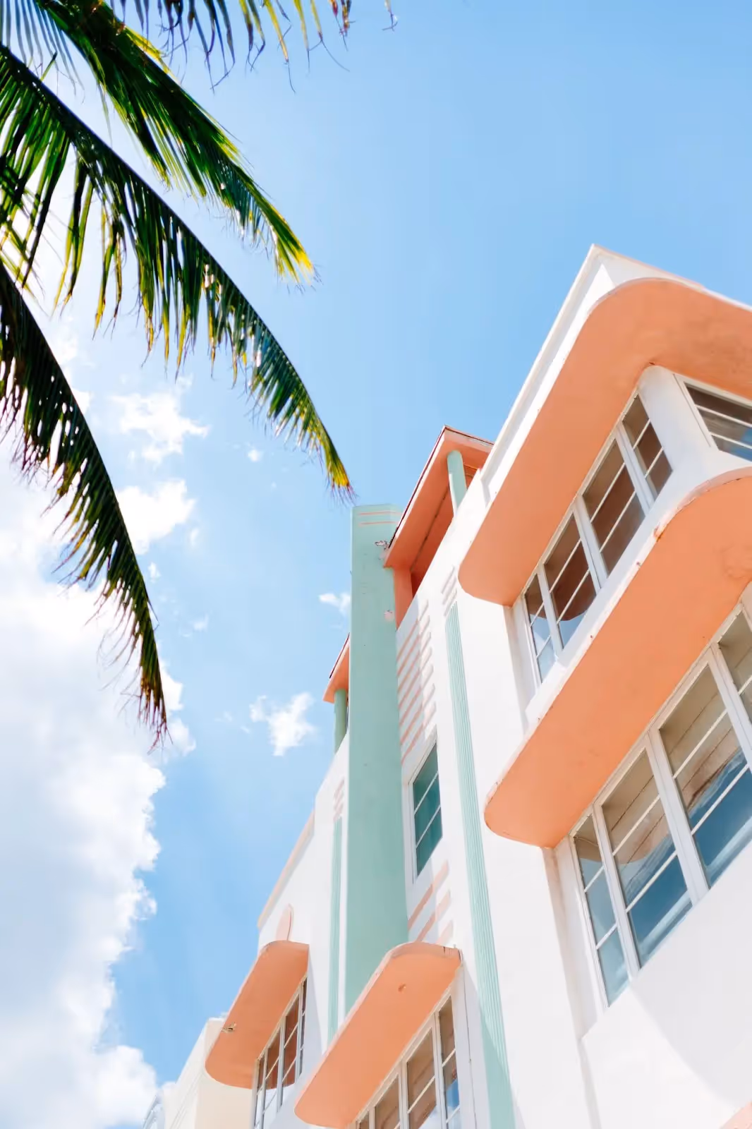 Art Deco building with peach balconies and mint accents under a blue sky, with palm fronds framing the top corner.