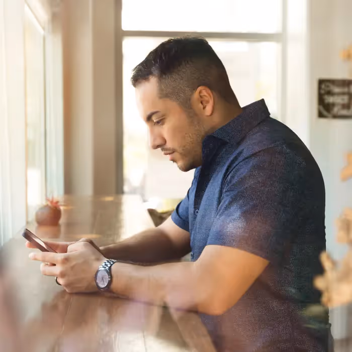 Man sitting by a window at a wooden counter, focused on reading a newsletter on his smartphone.