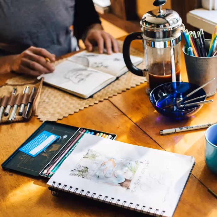 Artist sketching with coffee and stationery on wooden desk, illustrating creative email newsletter inspiration.