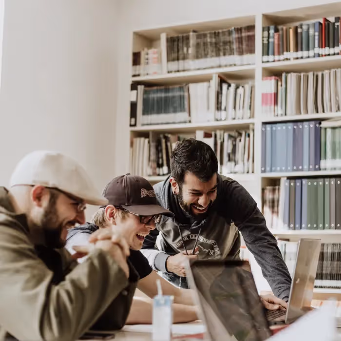 Group of young professionals laughing and collaborating around laptops in a library setting.
