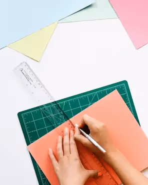 Paper crafting: top-down view of hands using an X-Acto knife and ruler to trim coral cardstock on a green mat with pastel sheets