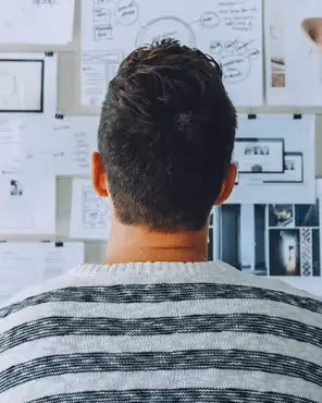 Man in striped sweater analyzing sketches, charts, and wireframes pinned on a creative planning wall.