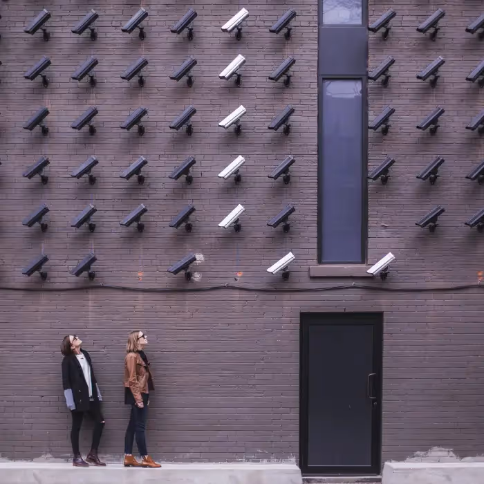 Two women standing under a wall covered with multiple security cameras, symbolizing privacy, data protection, and email compliance practices.