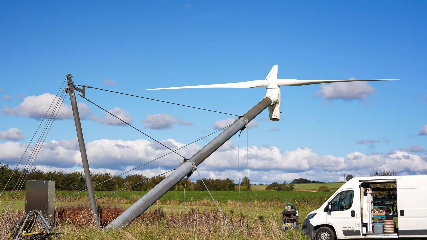 Partially assembled wind turbine with blades attached to a tilted tower supported by tension cables in a grassy field, next to a white van with open side door containing tools.