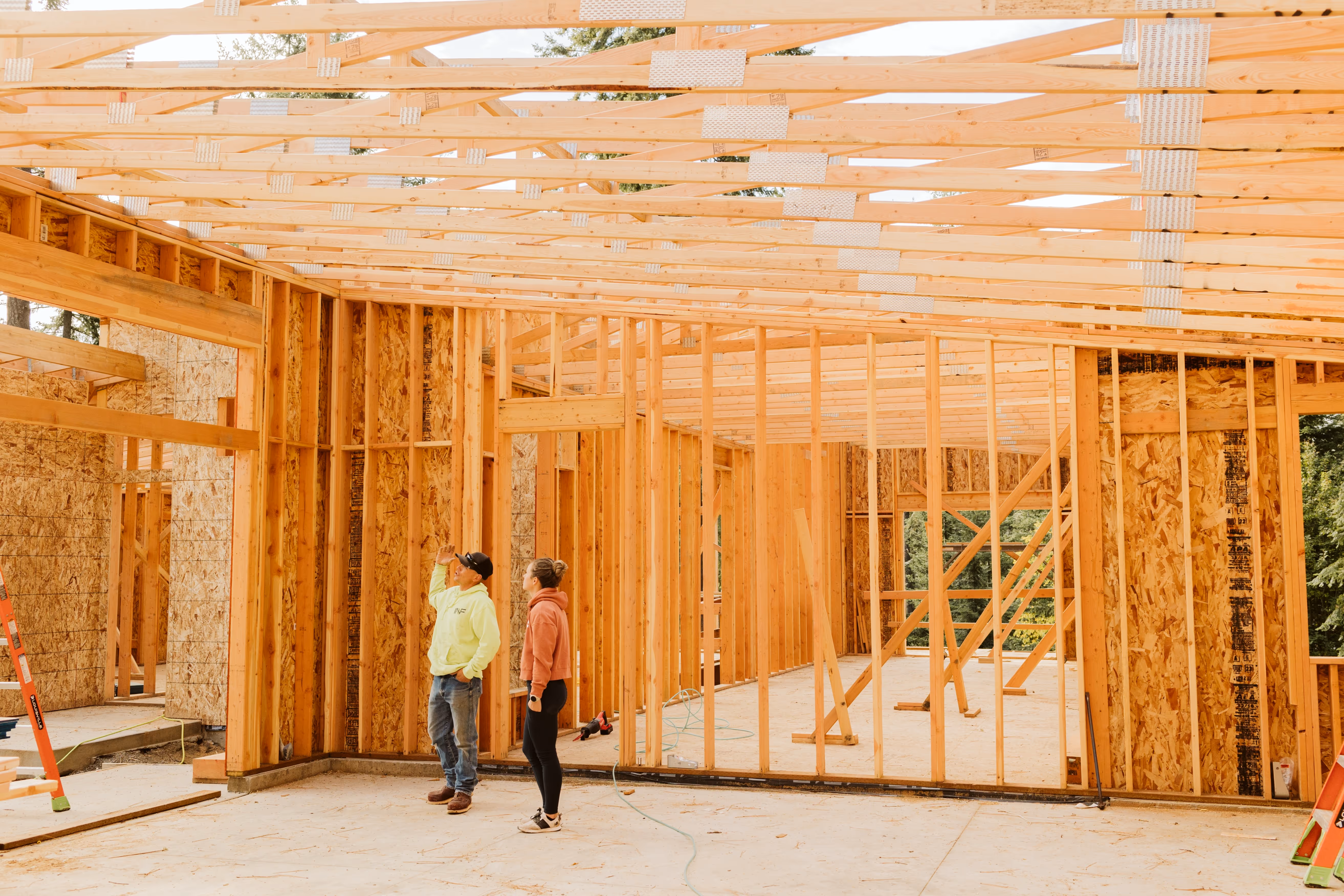 Two people inspecting wooden framing inside a house under construction with exposed ceiling beams and wall studs.