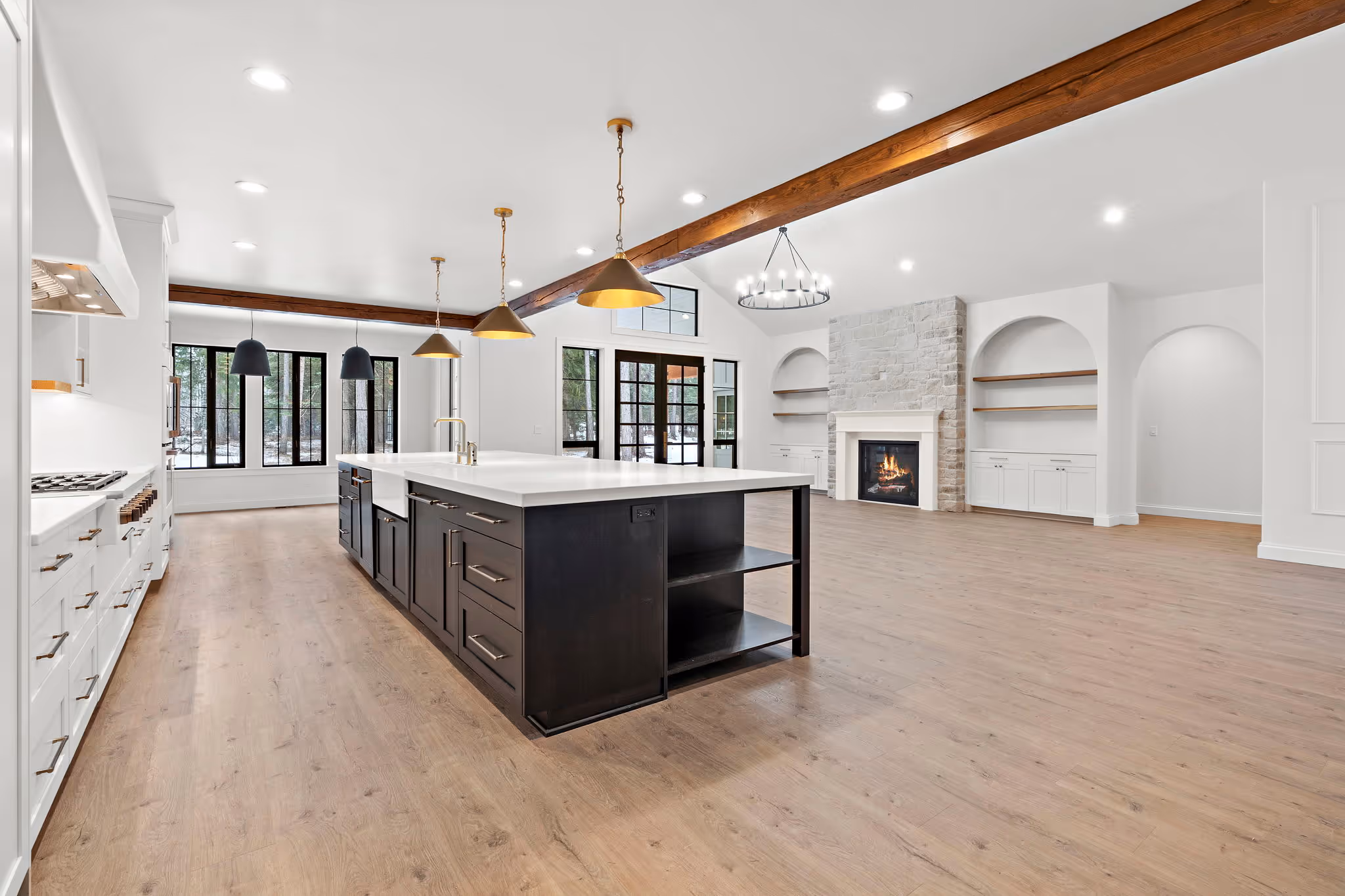 An open floor plan interior photo of a custom home. The home has VCC  floors, a gas fireplace, exposed wood beams, white quartz countertops, farmhouse sinks, and champagne bronze and matte black finishes. 