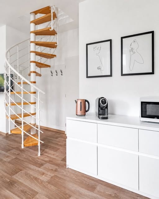 Moderní světlý interiér s bílou komodou v popředí, na které stojí rychlovarná konvice v barvě růžového zlata, kávovar a část mikrovlnné trouby. Nad komodou visí dva černě orámované obrazy s minimalistickými černobílými skicami ženských siluet. V pozadí vlevo se nachází točité schodiště s dřevěnými stupni a bílým kovovým zábradlím, vedoucí do patra. Podlaha má dekor dřeva.