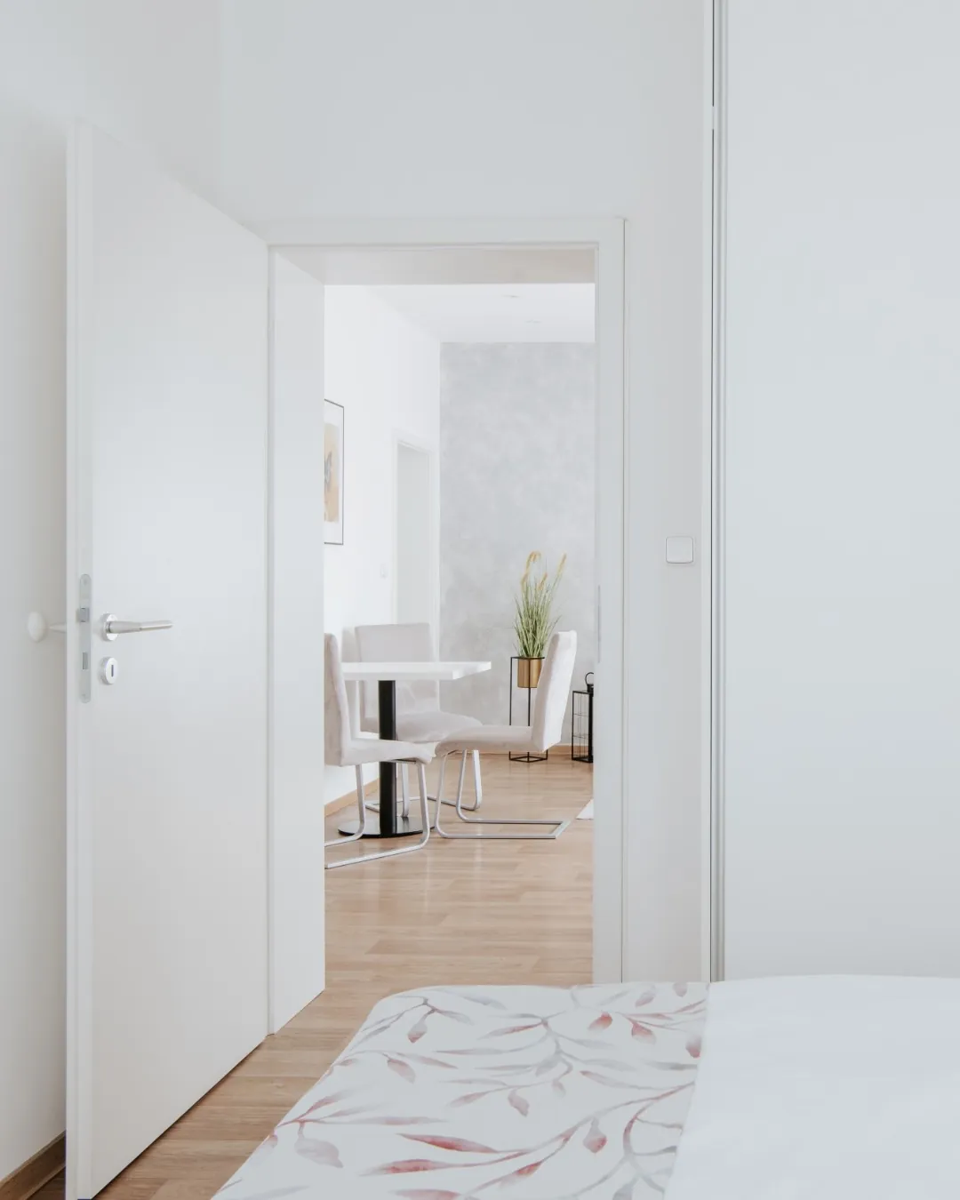 Pohled z ložnice apartmánu č. 4 přes otevřené bílé dveře do světlého obývacího pokoje s jídelním stolem a židlemi. V popředí okraj postele s přehozem s motivem listů.