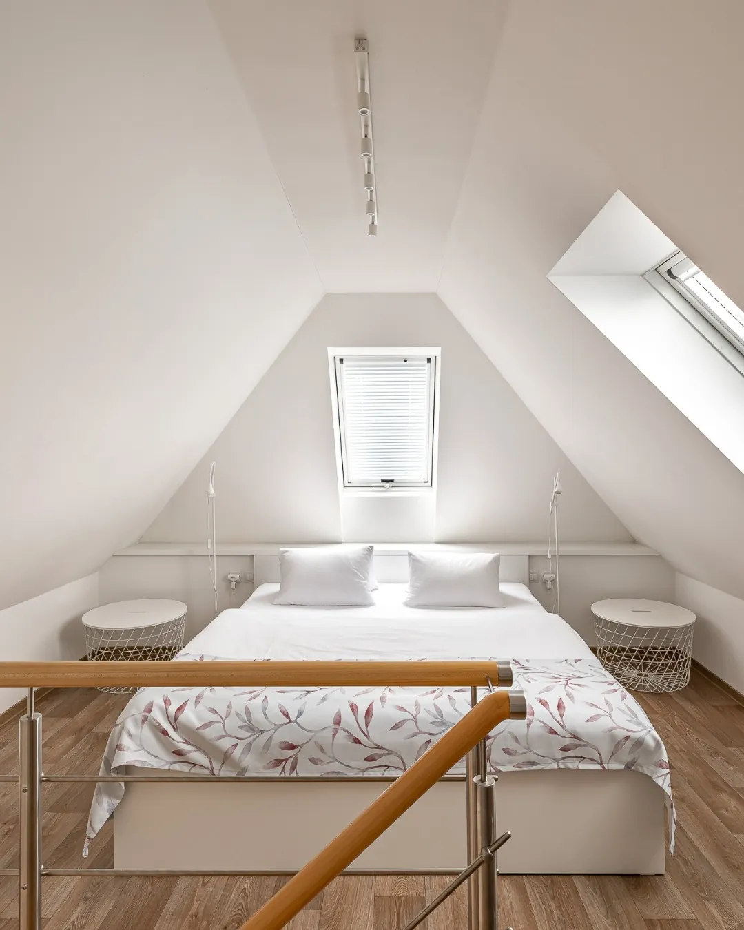 Podkrovní ložnice apartmánu č. 1 s manželskou postelí, bílými polštáři, přehozem s motivem listů a střešním oknem. V popředí dřevěné zábradlí galerie.