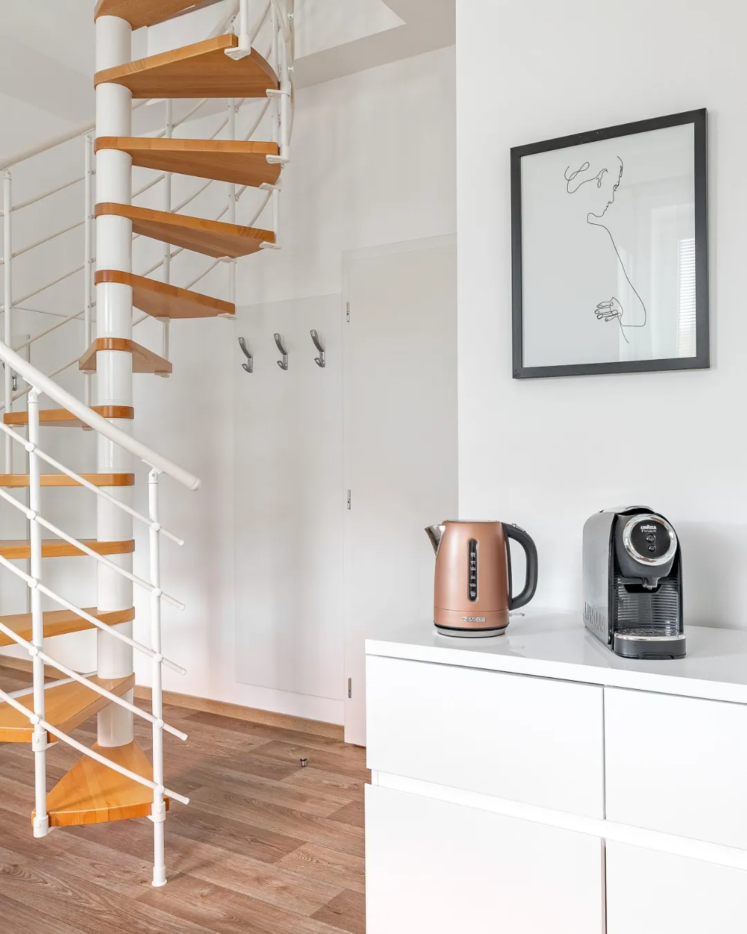 Točité schodiště s dřevěnými stupni a bílým zábradlím v mezonetovém apartmánu. Vedle stojí bílá komoda s rychlovarnou konvicí a kávovarem, na stěně visí obraz v černém rámu.