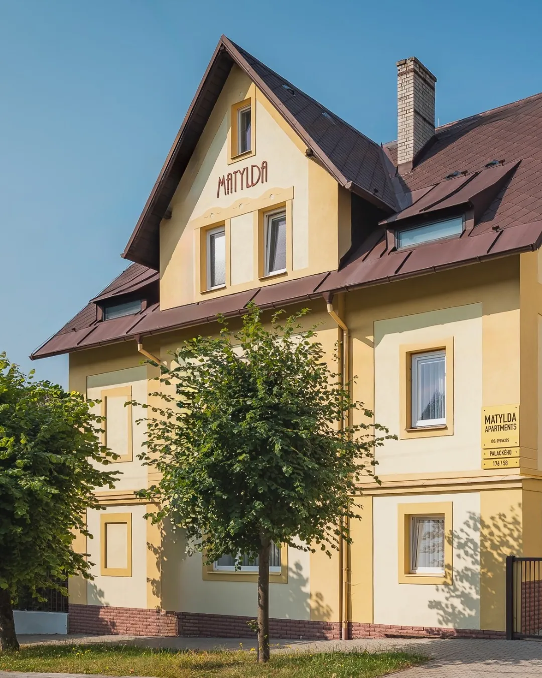 Žlutá vila Matylda s hnědou střechou, vikýři a nápisem Matylda na štítu. Před domem roste strom, nad vilou je modrá obloha.