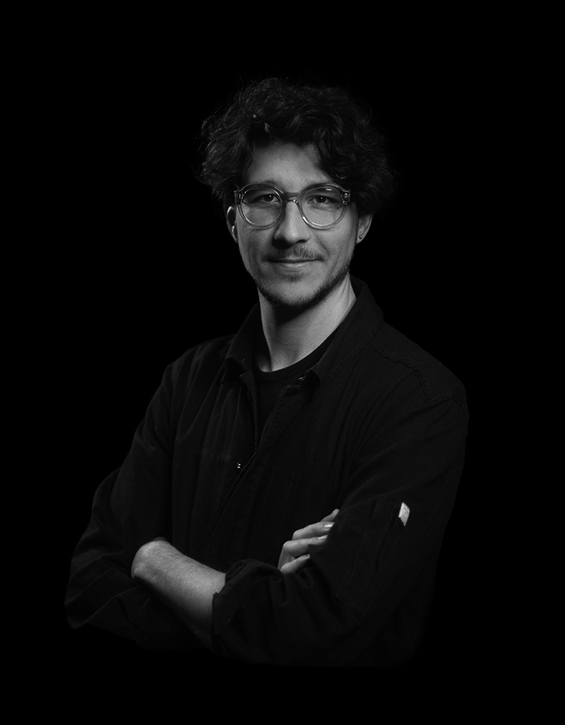 Young man with curly hair and glasses wearing a dark shirt, arms crossed, against a black background.