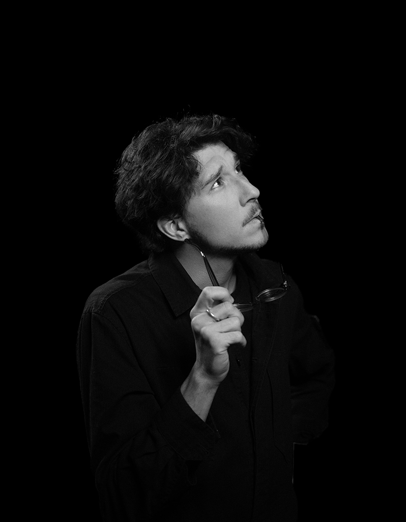 Black and white portrait of a young man with curly hair looking upward, holding glasses near his face.