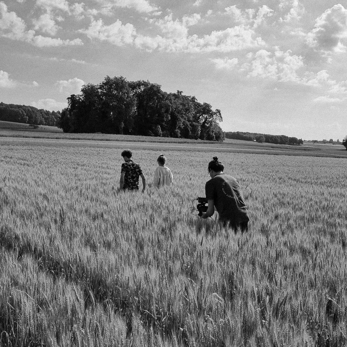 Three people walking through a wheat field with one person filming the others with a camera, under a partly cloudy sky.