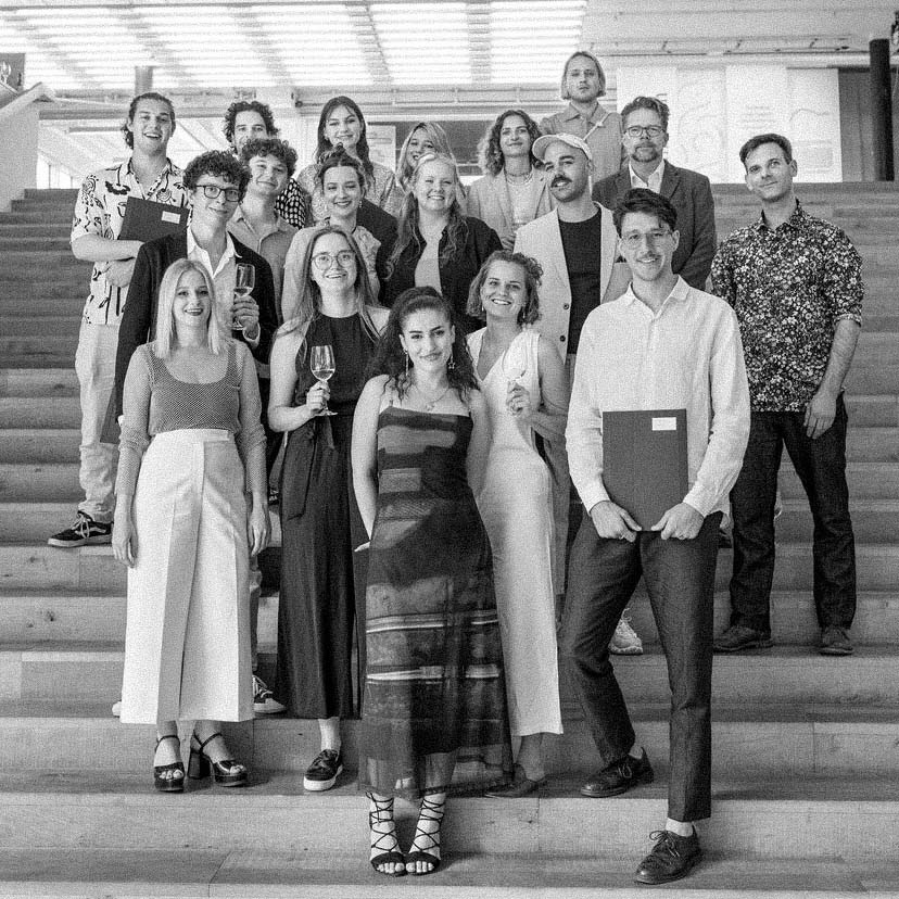 Group of eighteen young adults posing on indoor steps, some holding glasses and folders, dressed in semi-formal attire.