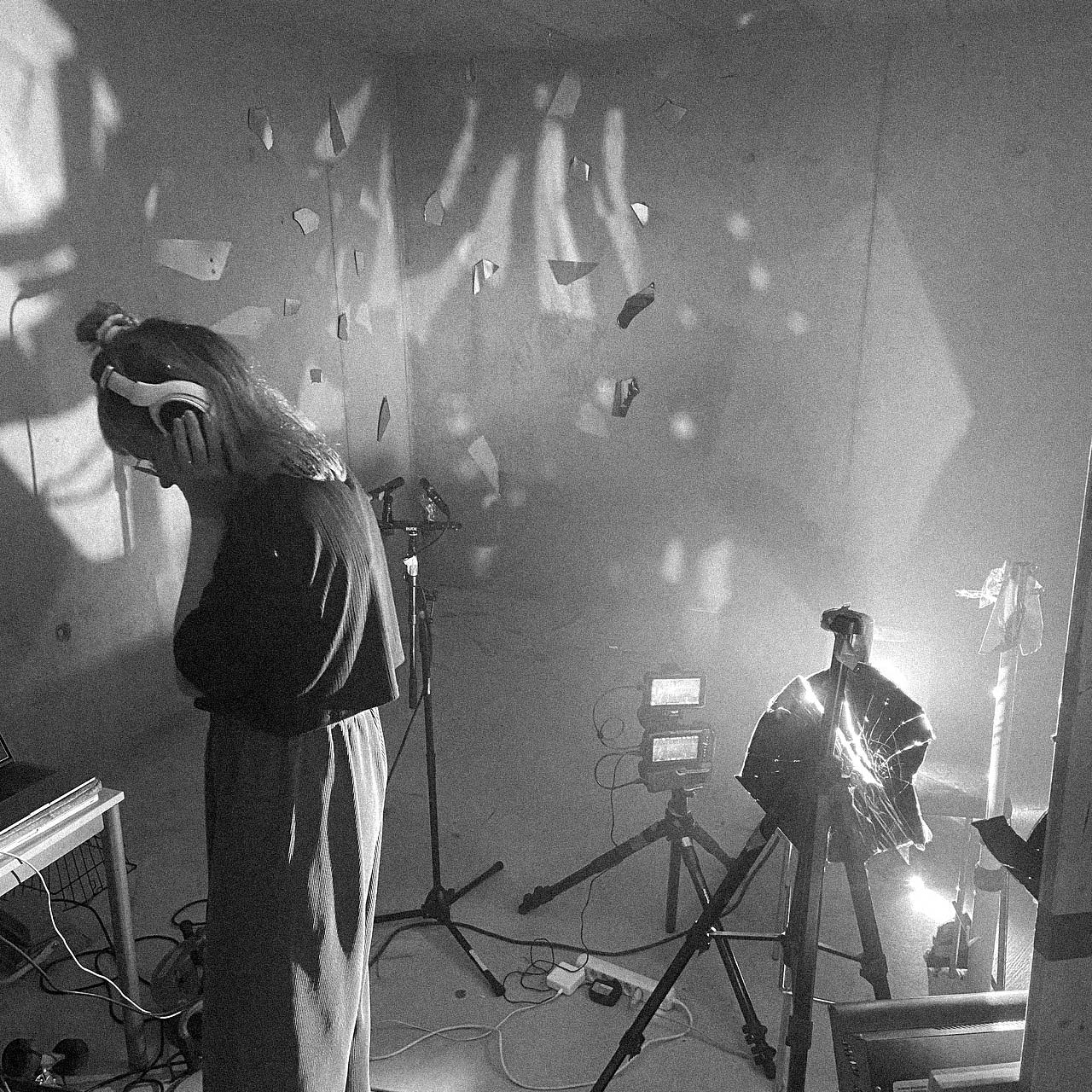 Black and white photo of a person wearing headphones and standing in a room with recording equipment and lights casting patterned shadows on the walls.