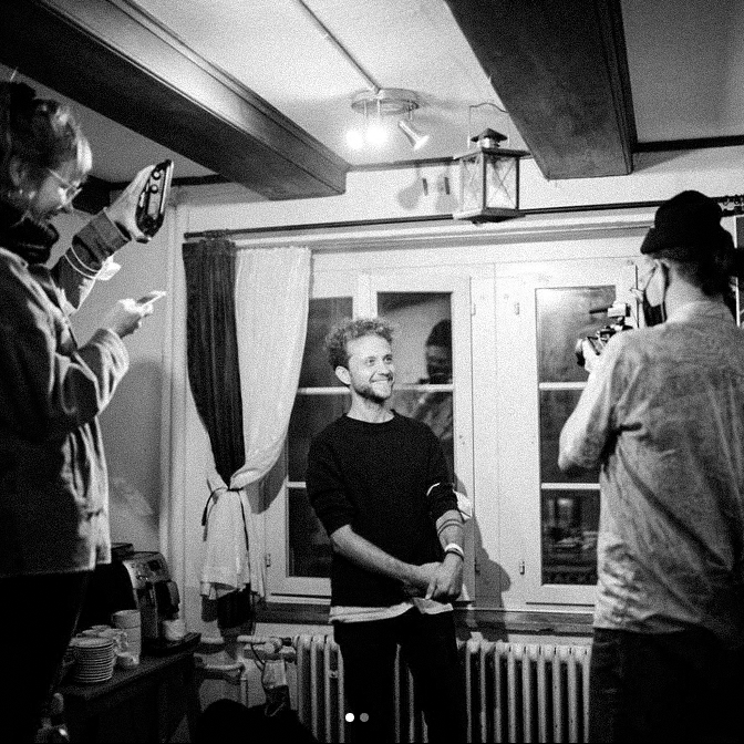 Black and white photo of a man smiling and rolling up his sleeve while two people film and photograph him indoors near a window.