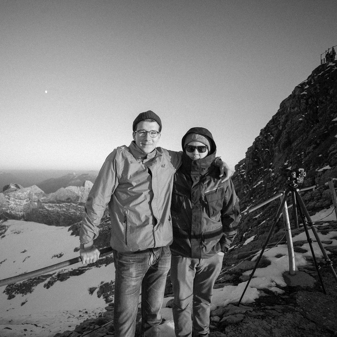 Two people dressed in winter jackets and hats stand arm in arm on a snowy mountain with rocky terrain at sunset.
