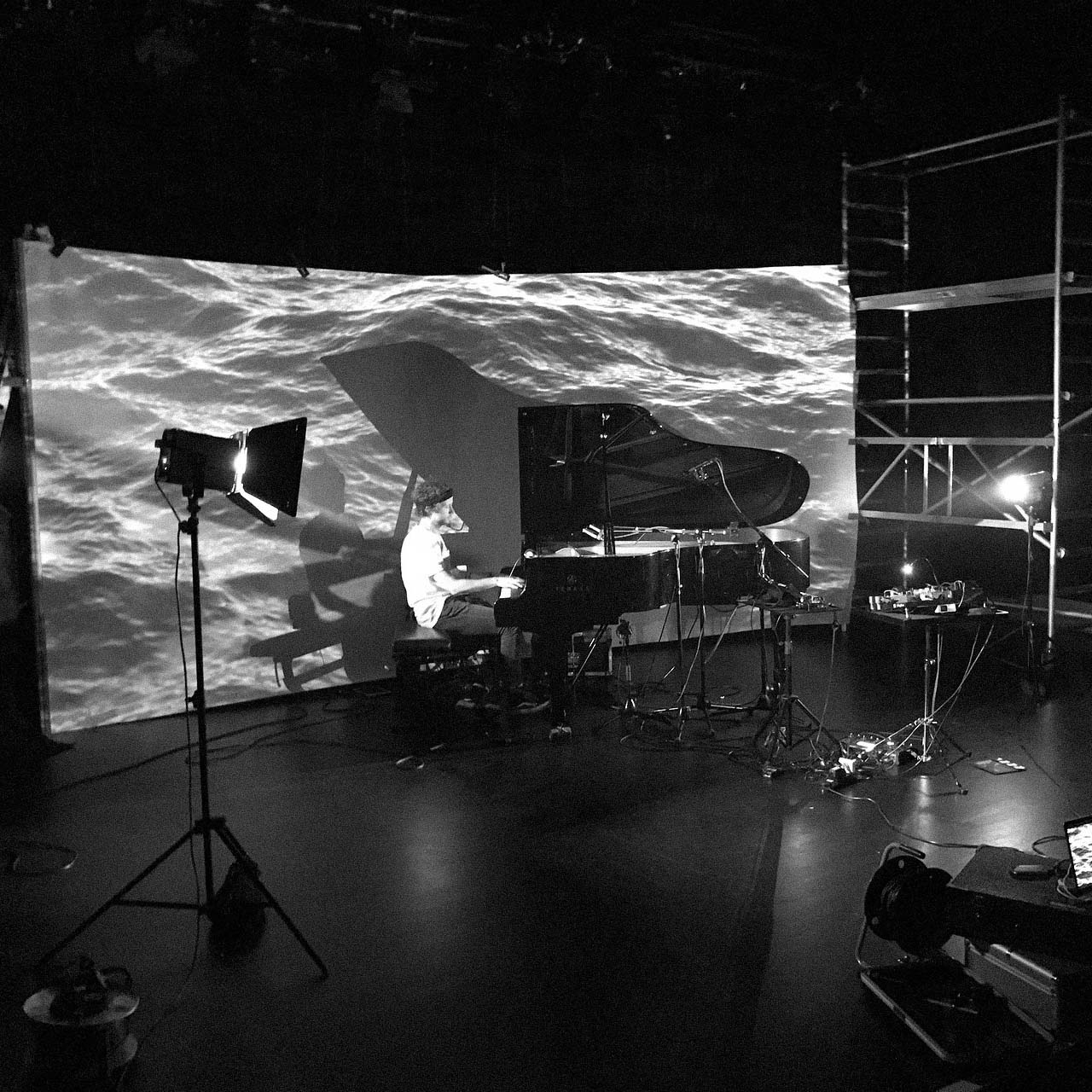Black and white image of a man playing a grand piano in a dark room with a water surface projection on the backdrop.