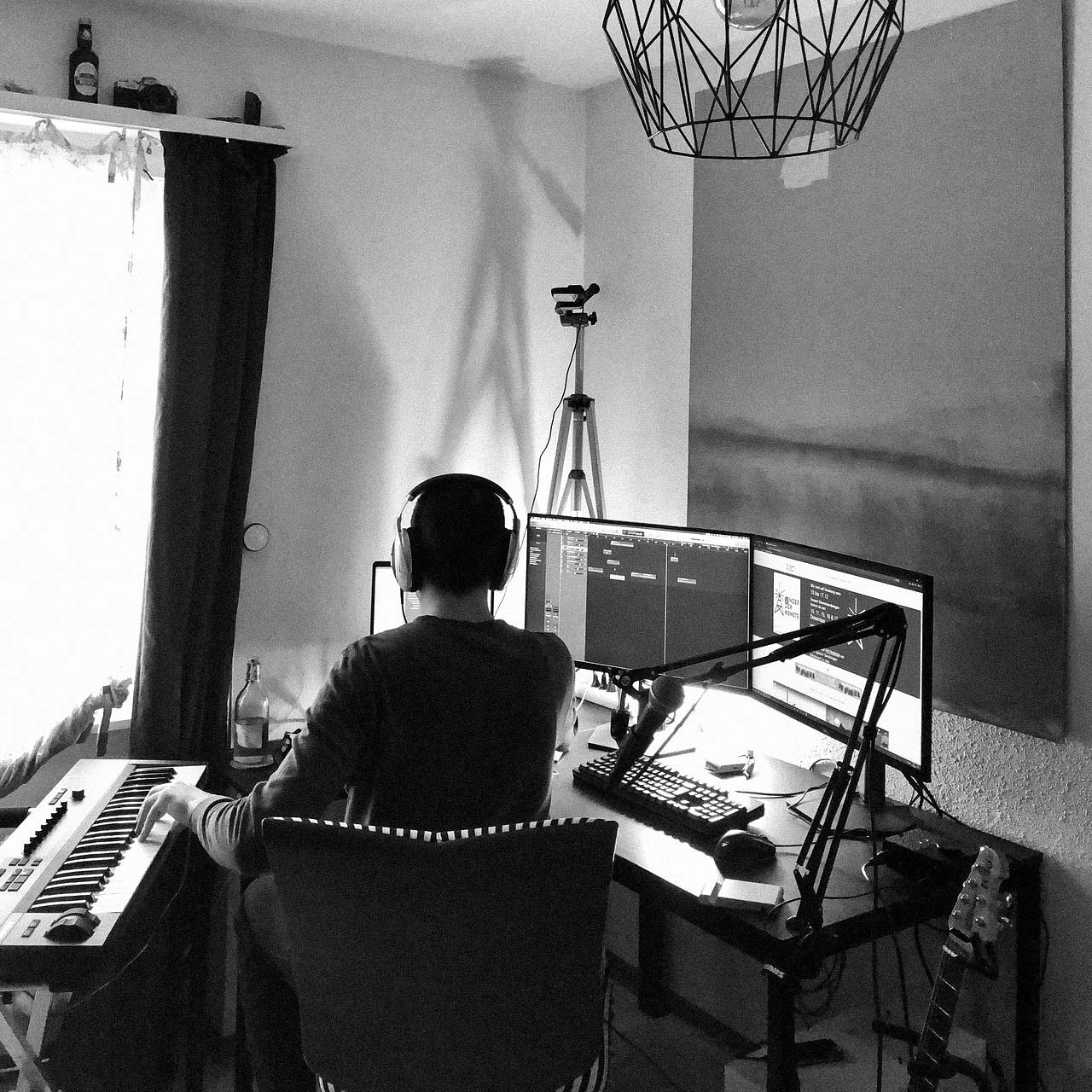 Person wearing headphones playing keyboard in a home studio with dual monitors, microphone, and guitar.