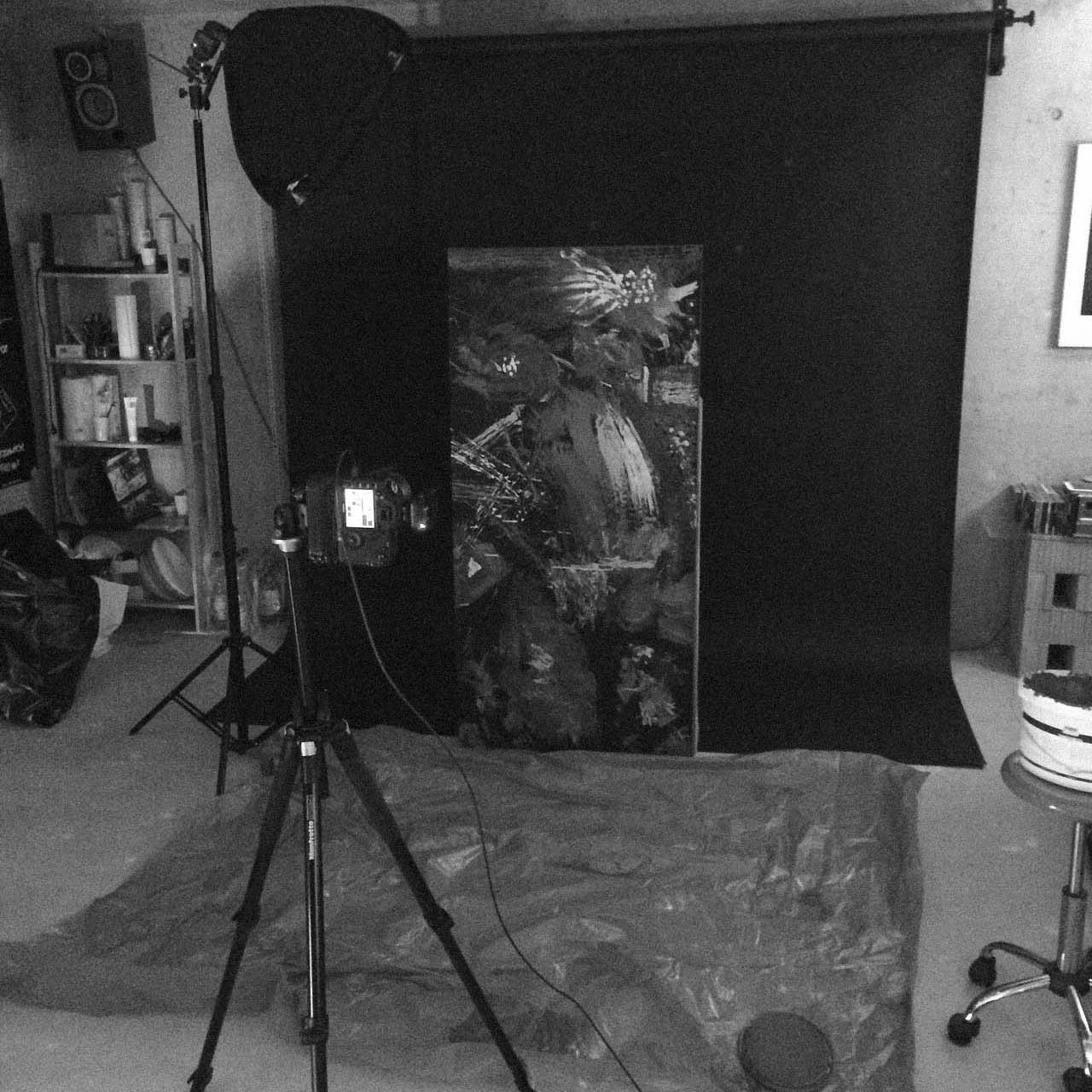 Photography setup in a studio with a camera on tripod facing an abstract painting against a black backdrop and a softbox light to the left.